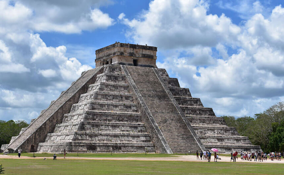 Two friends girls walking in direction to Chichen Itza pyramid while sun shines behind the building